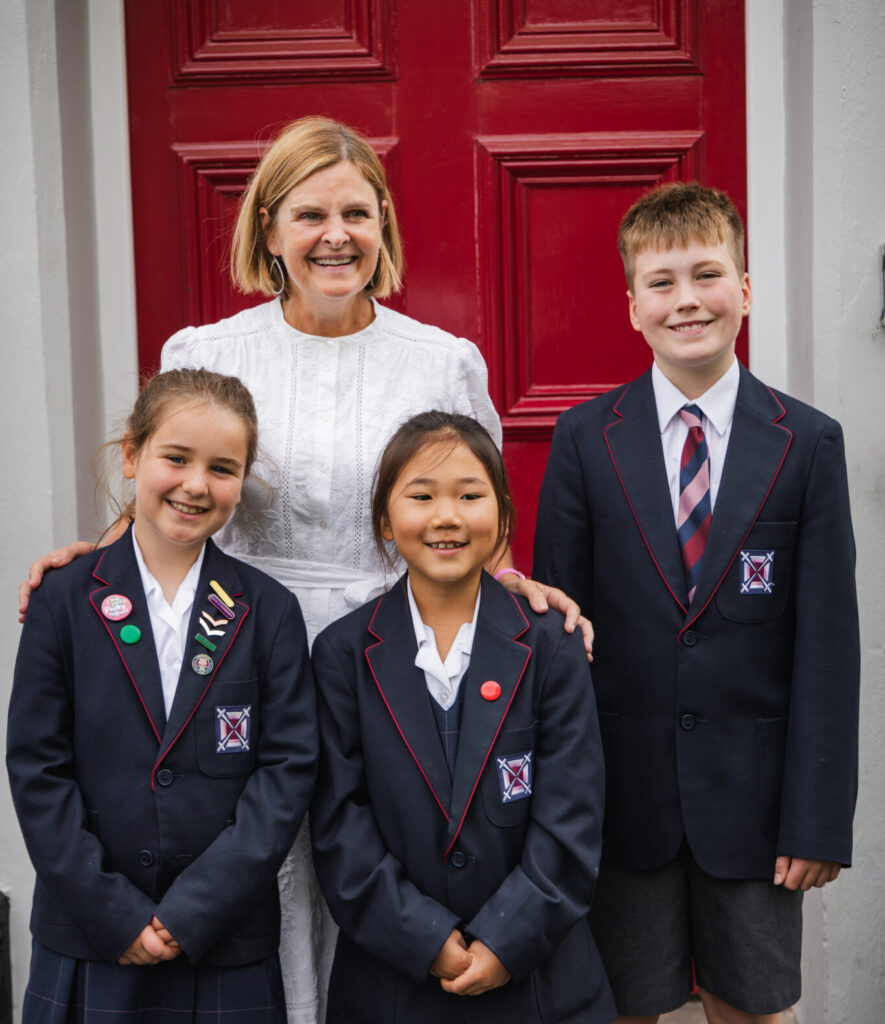 ES4 67 Copy – Exeter School Saskia in white dress with three junior pupils in smart uniform blazers in front of a red wooden door to the junior school