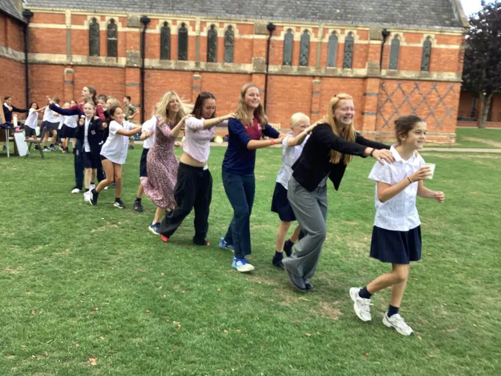 IMG 1186 – Exeter School Group of pupils and staff of all ages dancing in a line across a green field in front of red brick chapel with arms linked over shoulders