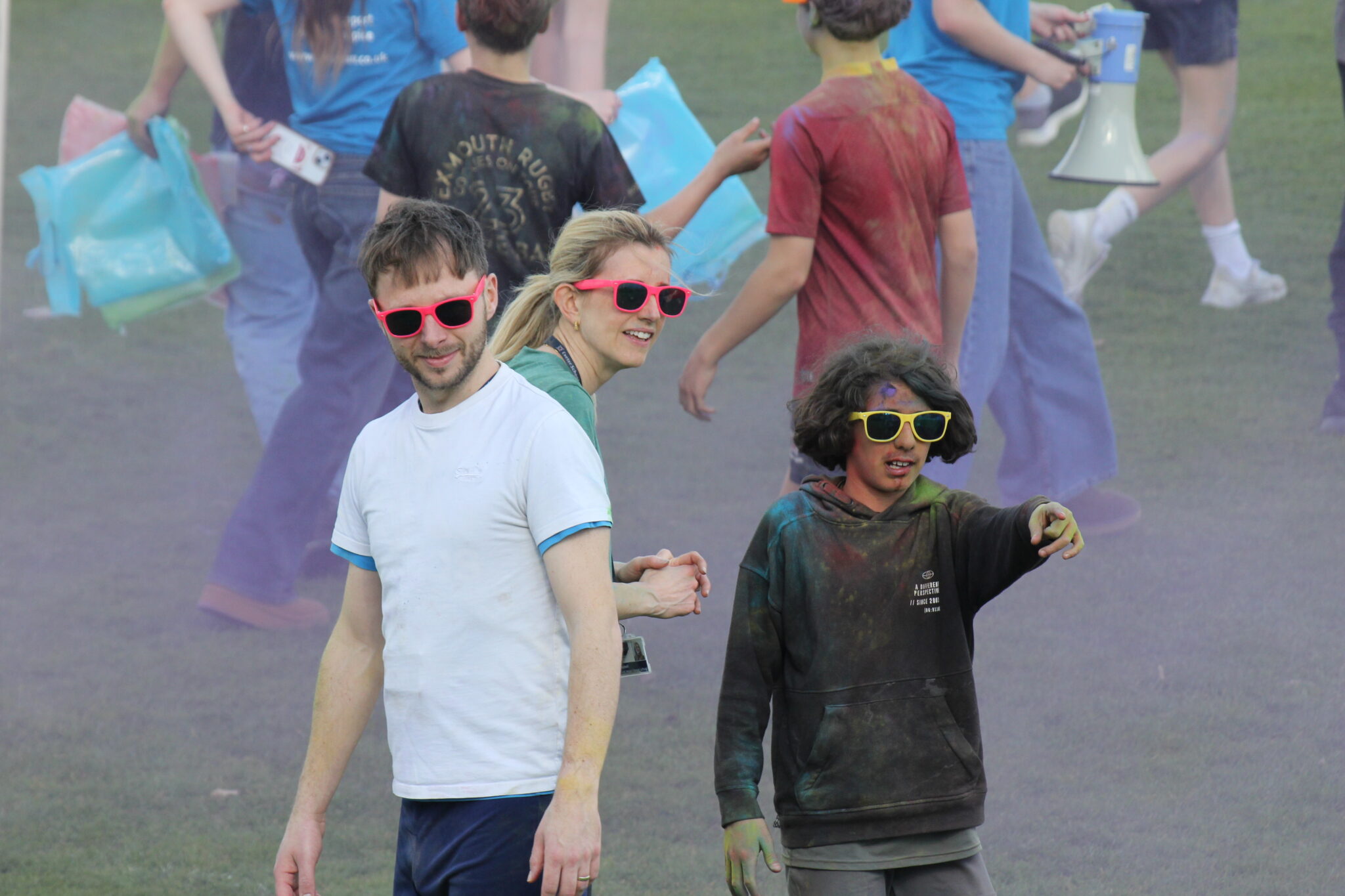 IMG 7565 – Exeter School young boy and two adults all in sunglasses on a field taking part in a colour fun run with paint powder clouds around them.