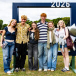 Six pupils, a mix of boys and girls, in non-school uniform, standing on the sports field in front of a large navy picture frame photo prop with balloons and 2025 numbers at the top
