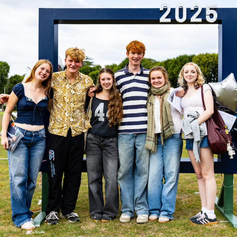 Six pupils, a mix of boys and girls, in non-school uniform, standing on the sports field in front of a large navy picture frame photo prop with balloons and 2025 numbers at the top