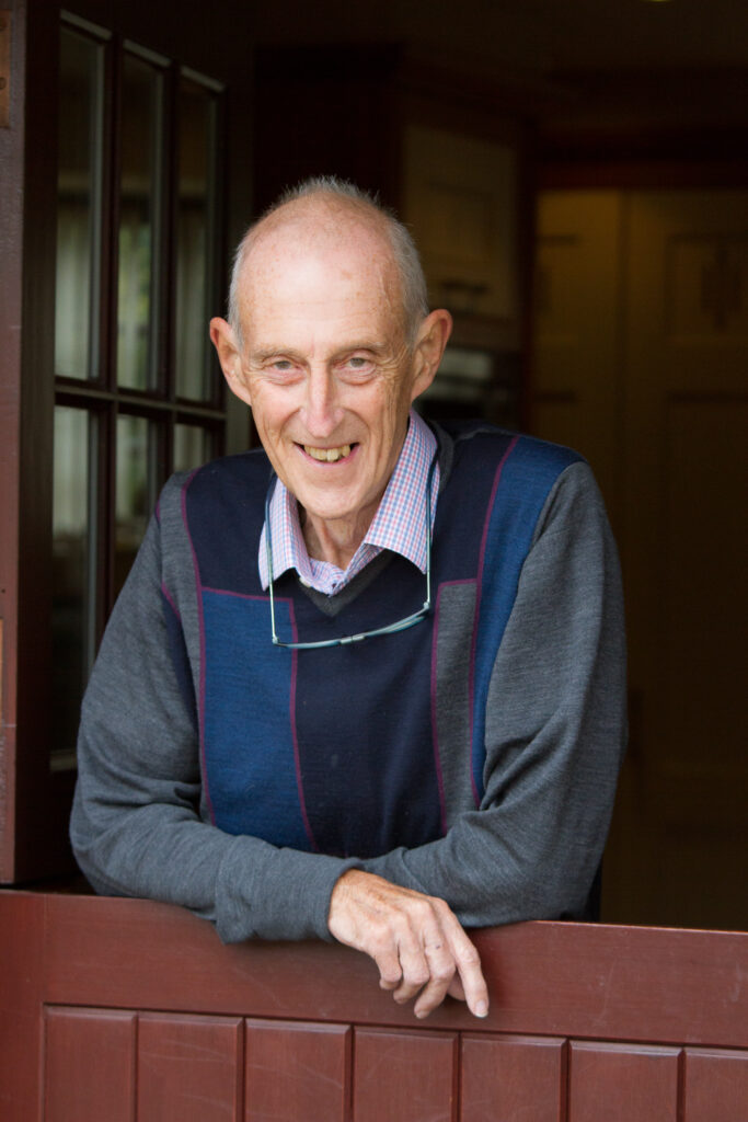 A happy older gentleman smiling at the camera leaning on a gate with glasses round his neck