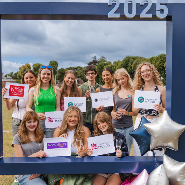 Group of young adults outside around a big picture frame photo prop holding cards stating their uni destinations