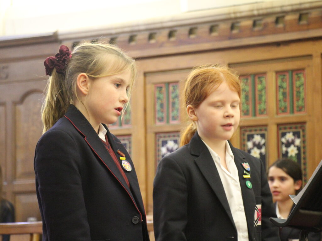 Two junior girl pupils singing side by side in a wood-panelled room.