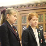 Two junior girl pupils singing side by side in a wood-panelled room.