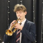 An Exeter School pupil in school uniform playing a trumpet on stage in front of a dark curtain backdrop.