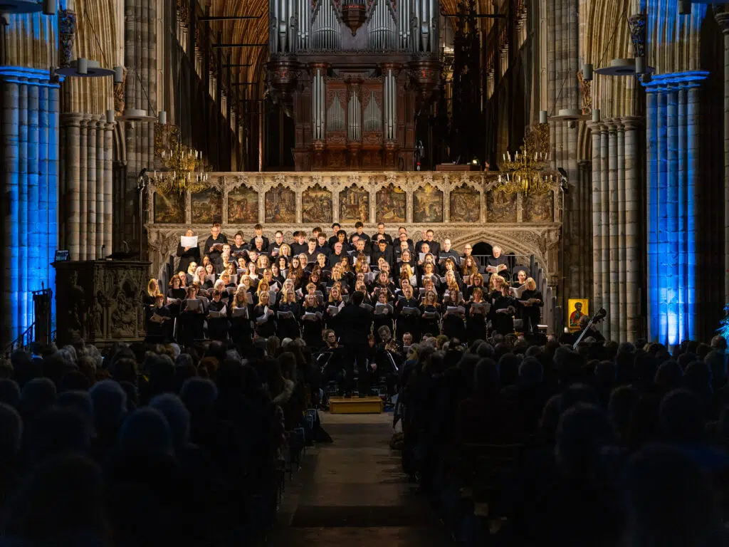A large school choir performing inside a cathedral, with a grand pipe organ and ornate stone architecture visible behind them.
