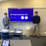 Two Exeter School students standing in front of a screen showing the IEA Budget Challenge 2026 presentation
