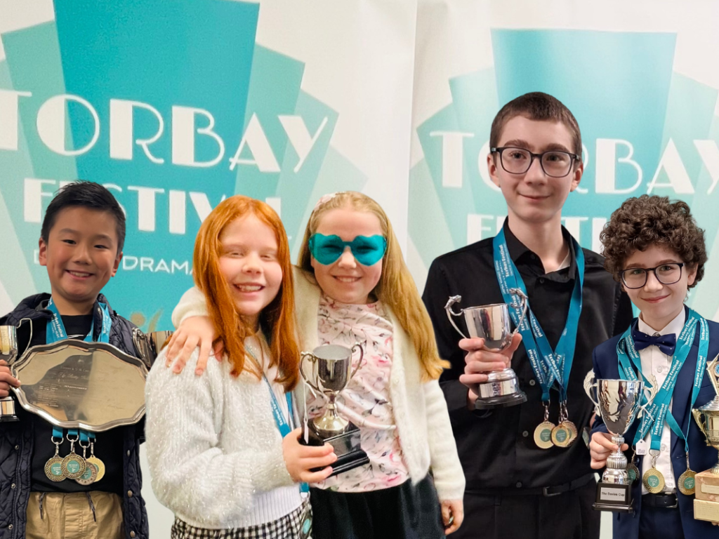 Pupils holding trophies and medals and smiling at the camera in front of a teal festival banner at the Torbay Music Festival.
