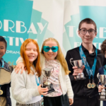 Pupils holding trophies and medals and smiling at the camera in front of a teal festival banner at the Torbay Music Festival.
