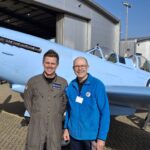 Two men stand smiling in front of a light-blue vintage aircraft outside a hangar.