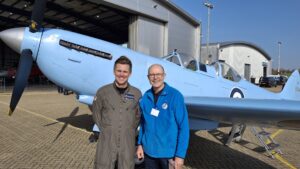 Two men stand smiling in front of a light-blue vintage aircraft outside a hangar.