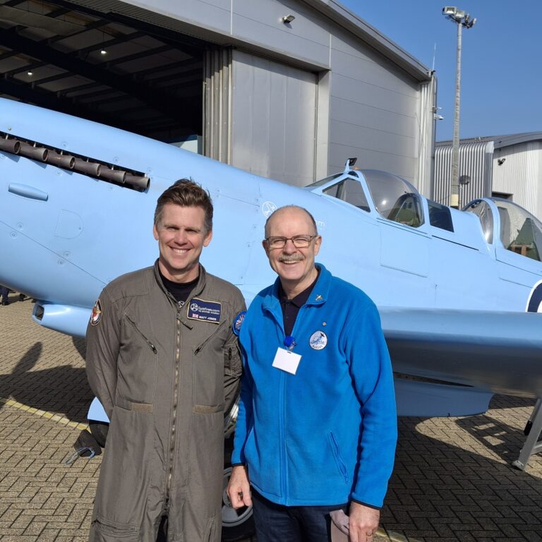 Two men stand smiling in front of a light-blue vintage aircraft outside a hangar.