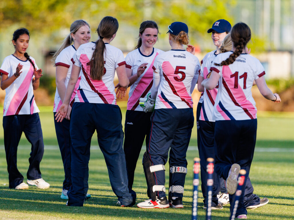 Group of female field hockey players in white jerseys with pink diagonal stripes, standing in a huddle on the field and chatting; visible numbers 5 and 11 on their backs.