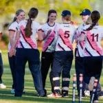 Group of female field hockey players in white jerseys with pink diagonal stripes, standing in a huddle on the field and chatting; visible numbers 5 and 11 on their backs.