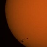 Close-up of the Sun's orange surface with a few tiny sunspots near the lower left edge.