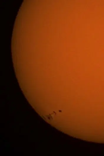 Close-up of the Sun's orange surface with a few tiny sunspots near the lower left edge.