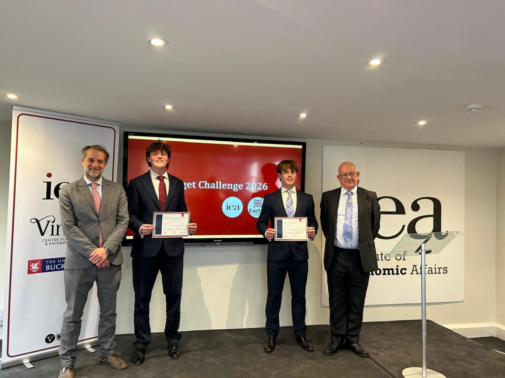Four men in suits pose on a stage, two young recipients hold certificates, flanked by two older men.