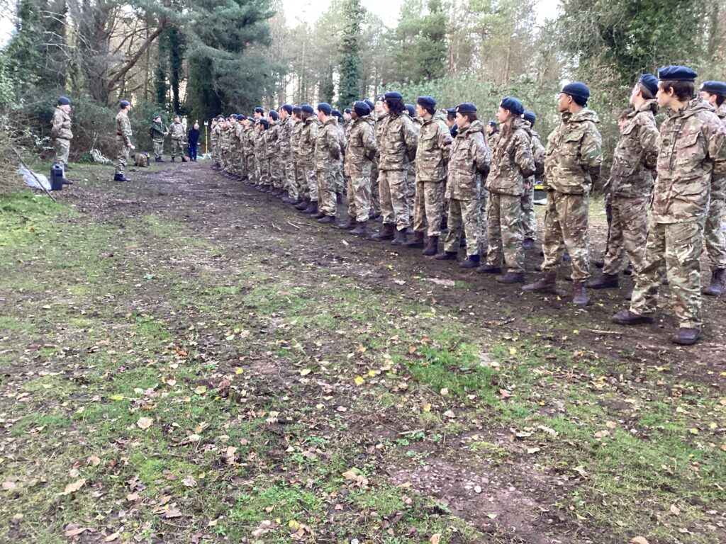 Exeter School CCF cadets in camouflage uniform and blue berets standing in a long formation in a woodland outdoor setting