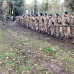 Exeter School CCF cadets in camouflage uniform and blue berets standing in a long formation in a woodland outdoor setting