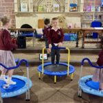 Children in maroon school uniforms bounce on small blue trampolines in a library-like room with bookshelves behind them.