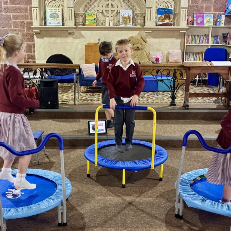 Children in maroon school uniforms bounce on small blue trampolines in a library-like room with bookshelves behind them.
