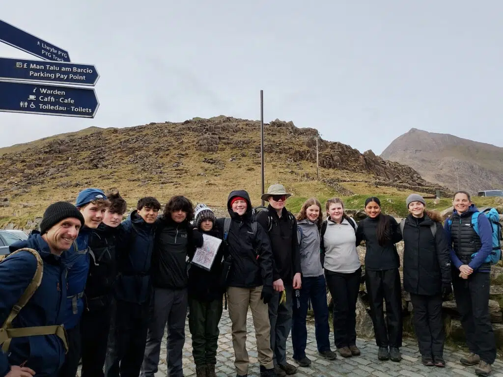 Group of hikers posing together on a cobblestone path beside blue signposts, rocky hillside behind them and a mountain in the distance.