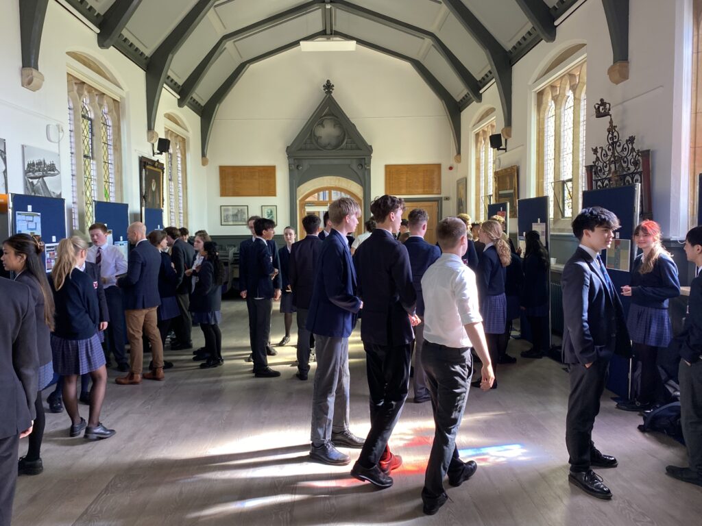 Exeter School pupils and adults mingling during an event in a grand school hall with vaulted ceilings and tall stained glass windows.
