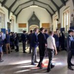 Exeter School pupils and adults mingling during an event in a grand school hall with vaulted ceilings and tall stained glass windows.