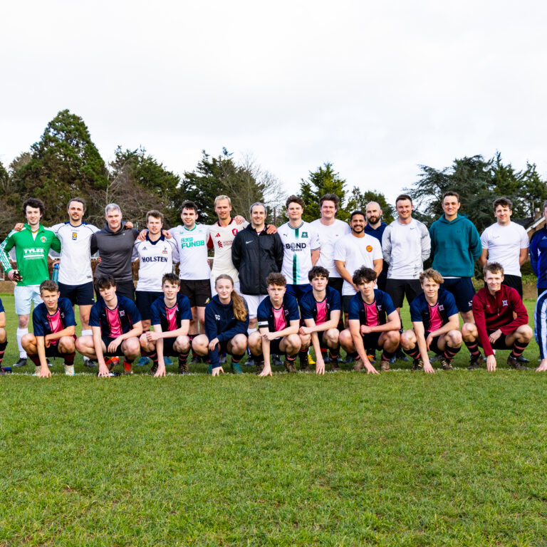 A large group of rugby players pose for a team photo on a grassy field, some kneeling in front and others standing, trees in the background.
