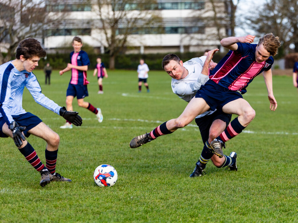 Soccer players chase the ball on a grassy field during a match, one in a light-blue kit sliding toward the ball while others run nearby.