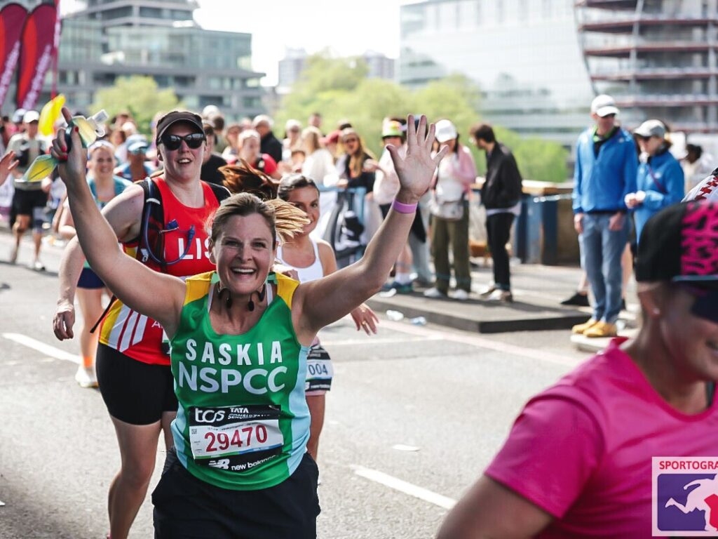 Smiling female runner in a green Saskia NSPCC jersey raises her arms as a cheering crowd lines a city street race behind her.