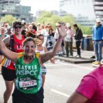 Smiling female runner in a green Saskia NSPCC jersey raises her arms as a cheering crowd lines a city street race behind her.