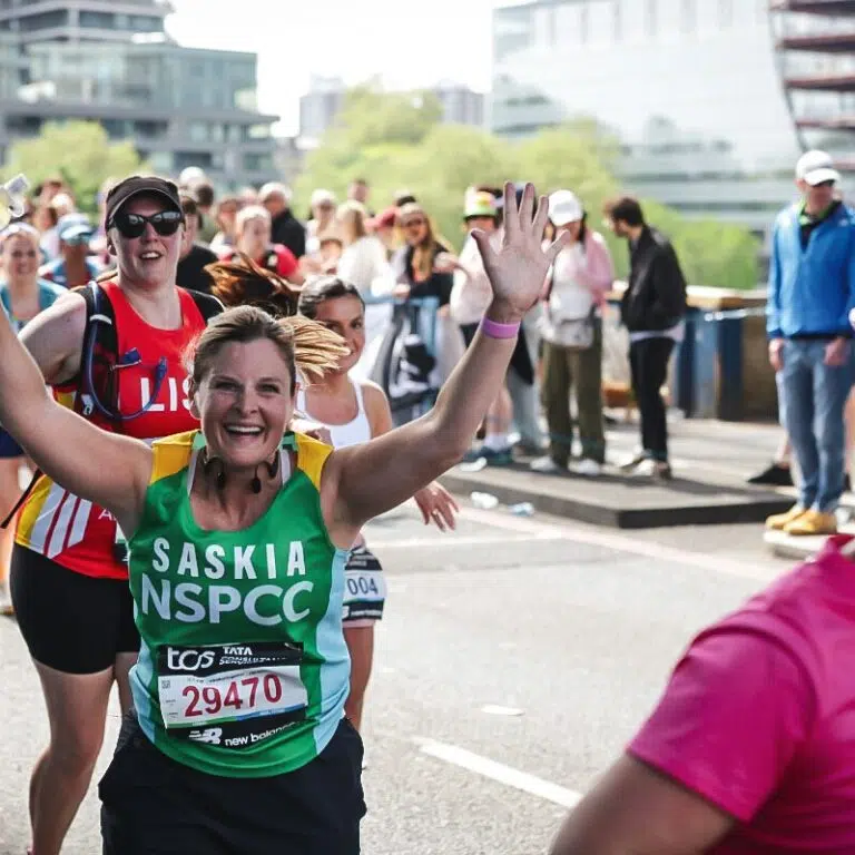 Smiling female runner in a green Saskia NSPCC jersey raises her arms as a cheering crowd lines a city street race behind her.