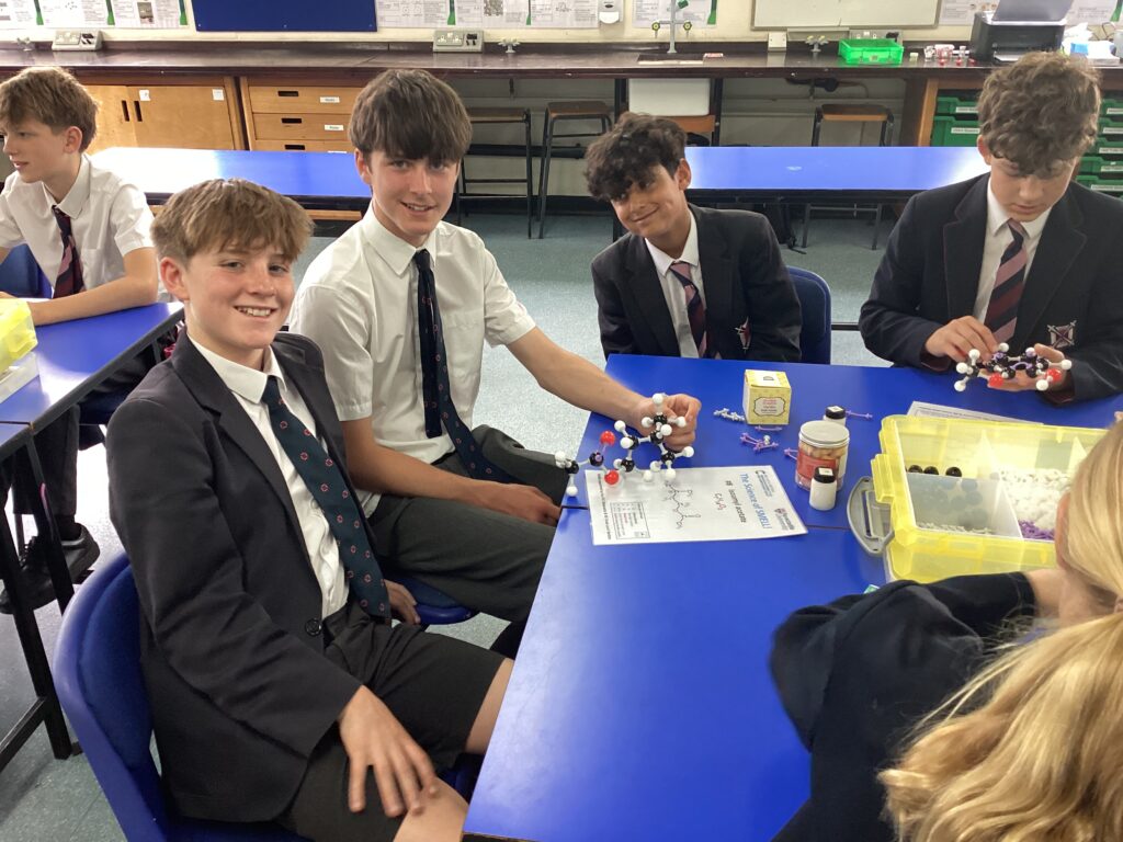 Students in school uniforms work at a lab table, building molecular models with plastic atoms in a science class setting.
