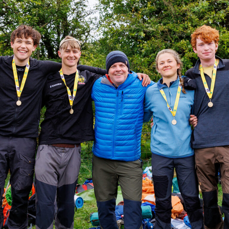 Seven young people with medals around their necks, standing with arms around each other outdoors on a grassy field.