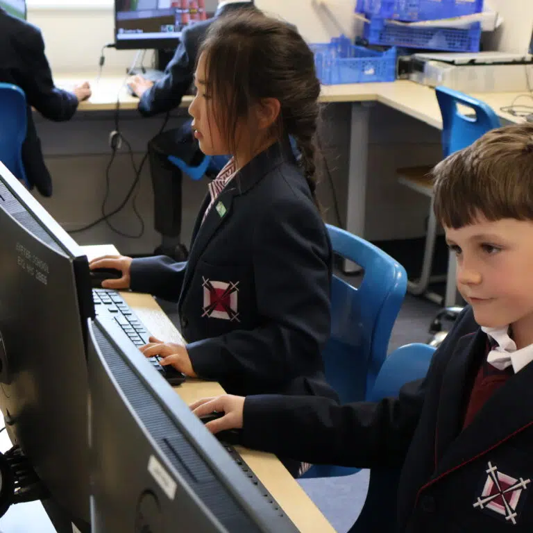 Two students in uniform sit at desktop computers in a classroom, focused on the screens.