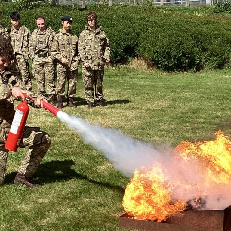 Soldiers in camouflage watch as a trainee uses a fire extinguisher on a controlled fire.