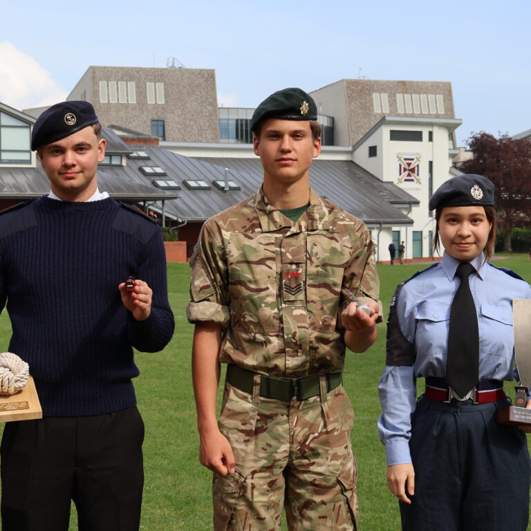 Three young cadets in uniform pose outdoors on a grassy area, holding awards with buildings in the background.
