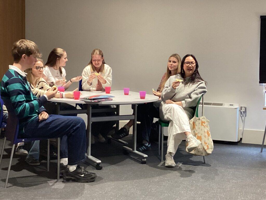 Group of young people sitting around a round table, eating snacks in a classroom or meeting room.