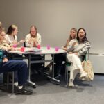 Group of young people sitting around a round table, eating snacks in a classroom or meeting room.