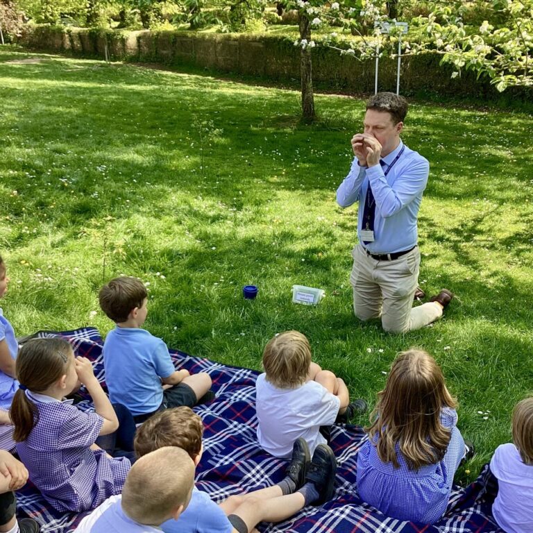 A group of children sits on a plaid blanket in a sunny park, listening to a man kneeling as he presents outdoors.