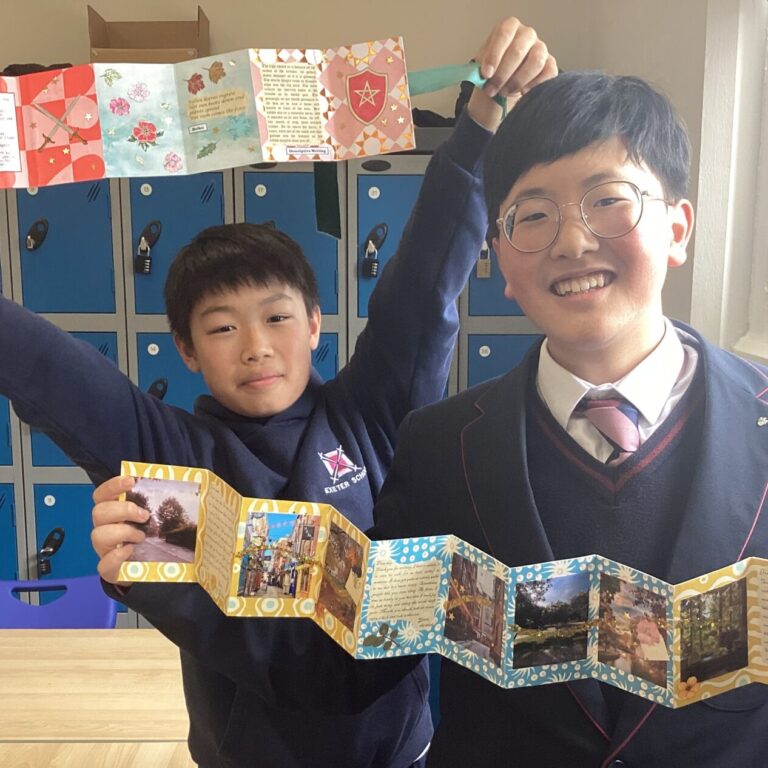 Two students in uniform proudly hold and display a long, colorful photo book in a classroom with blue lockers in the background, smiling at the camera.