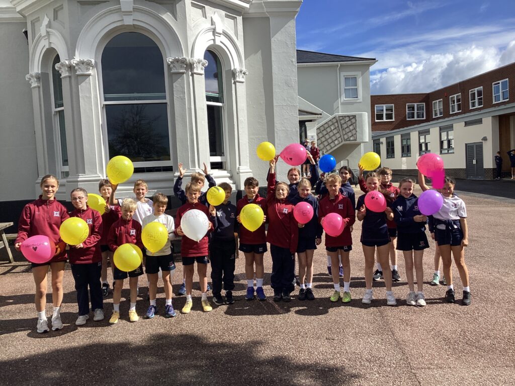Group of smiling schoolchildren in matching uniforms holding colorful balloons outside a white building with arched windows.