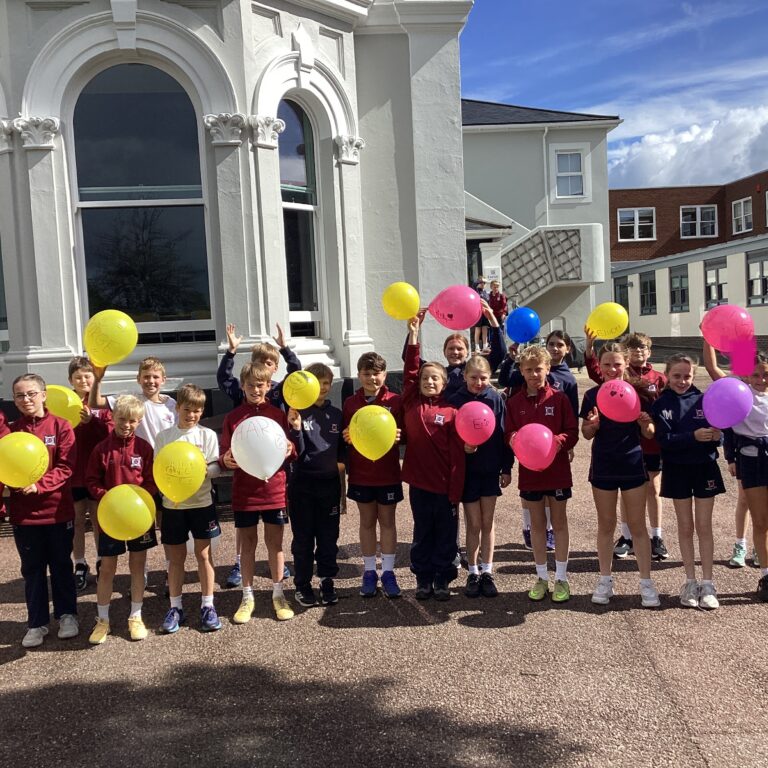 Group of smiling schoolchildren in matching uniforms holding colorful balloons outside a white building with arched windows.