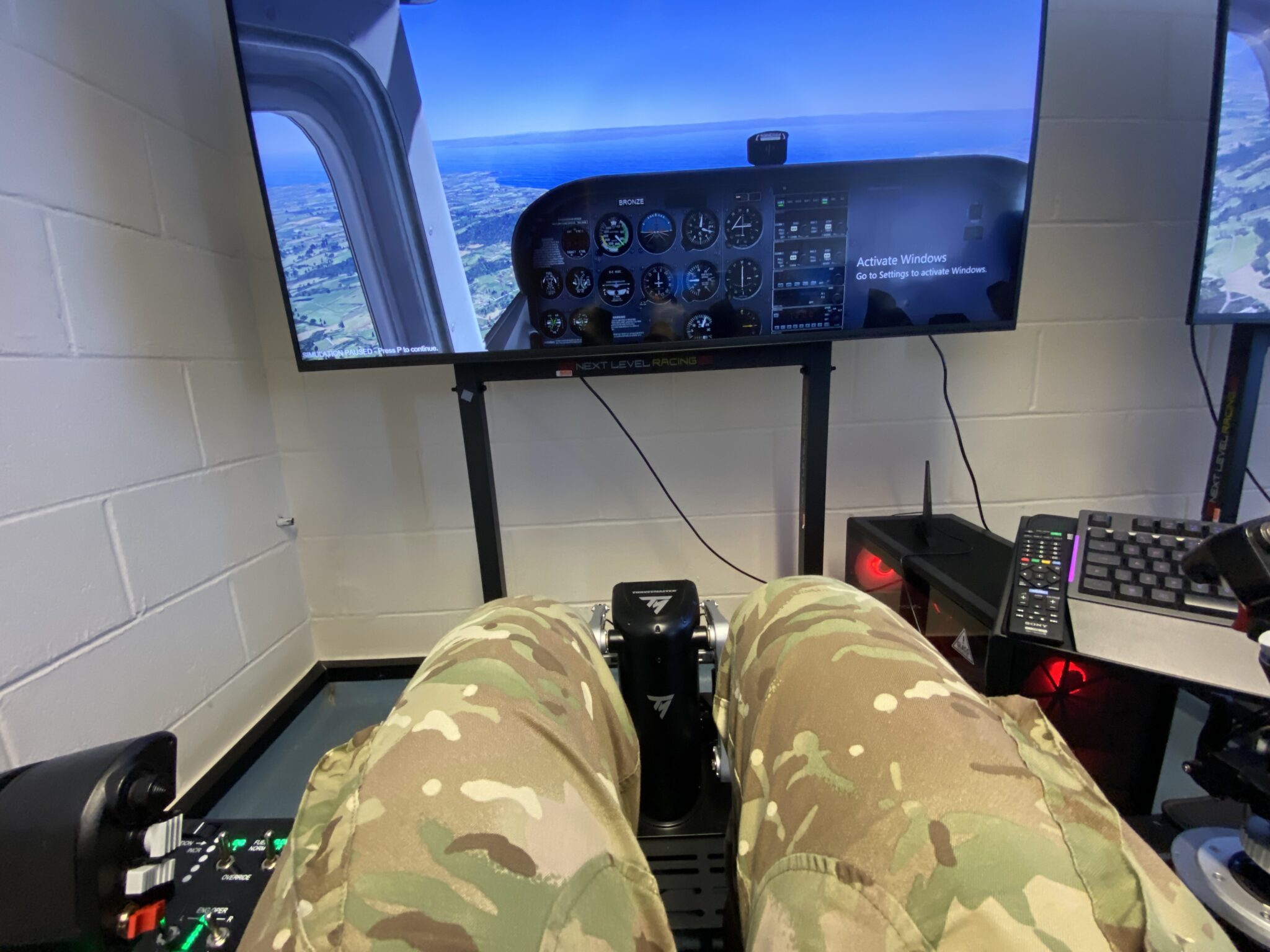 Person in camouflage pants seated in a flight-simulator cockpit, facing a large monitor showing an aircraft instrument panel and a landscape view outside the window.