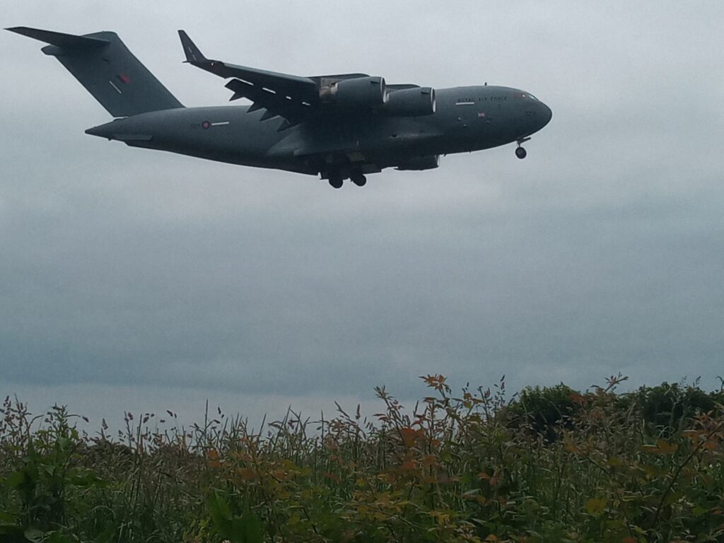 Grey four-engine military transport aircraft on final approach with landing gear down over a grassy field and shrubs under a cloudy sky.