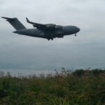 Grey four-engine military transport aircraft on final approach with landing gear down over a grassy field and shrubs under a cloudy sky.