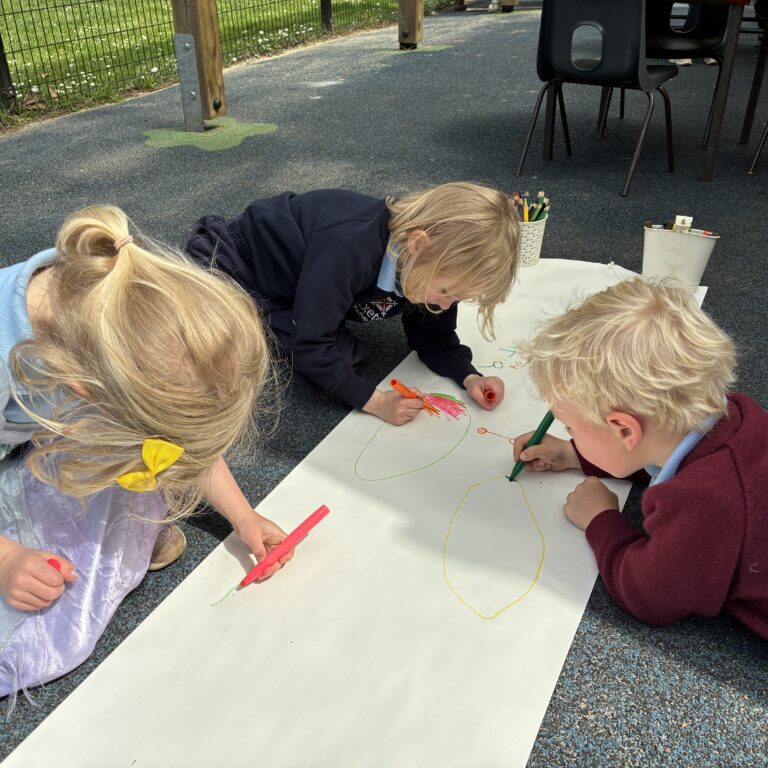 young children in uniform with their heads down drawing on a long roll of paper outside in the playground