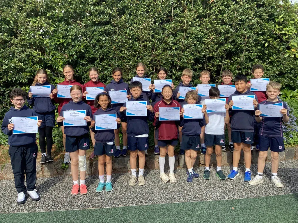 Group of children holding certificates outdoors in front of a green hedge, smiling at the camera.